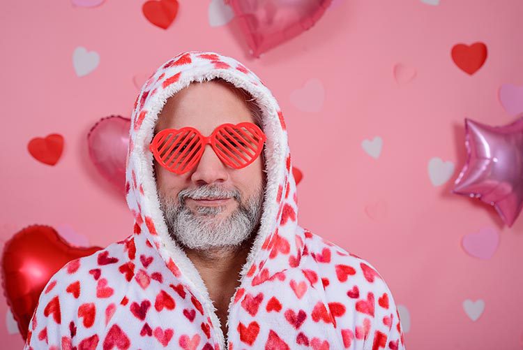 Valentine's Day portrait of gray-haired man wearing heart-shaped glasses and bathrobe with red hearts. Pink background with balloons.