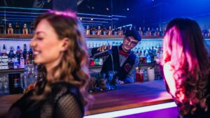 Man mixing a cocktail at a bar with two women in the foreground