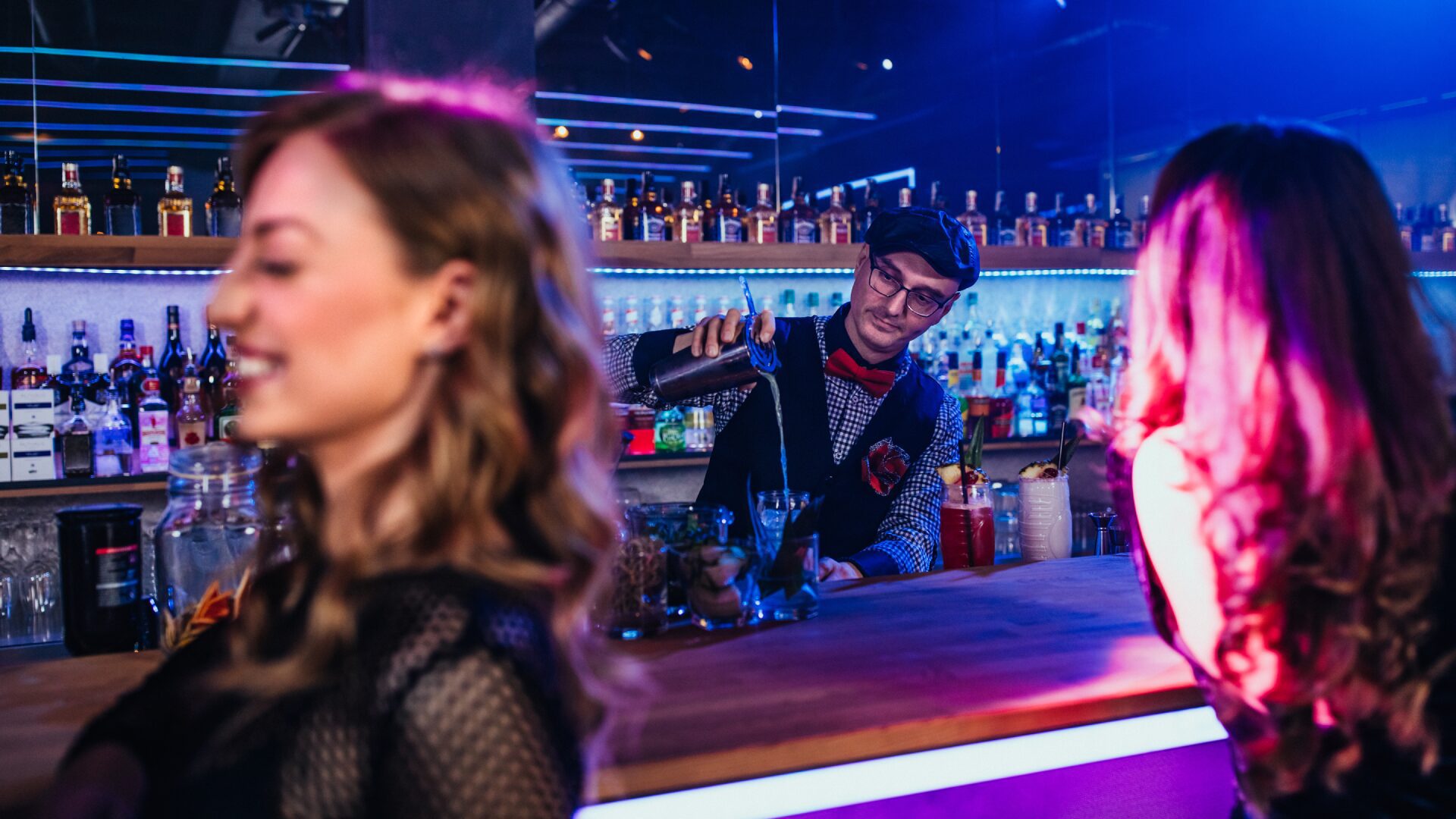 Man mixing a cocktail at a bar with two women in the foreground